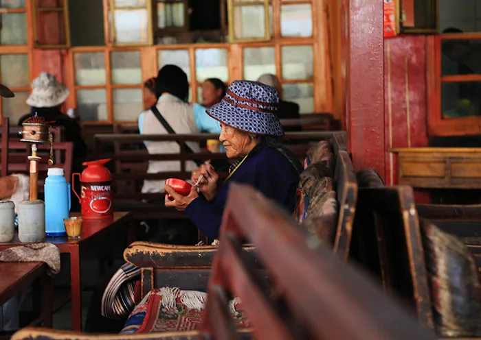 An elderly woman sits at a wooden table beside a mani prayer wheel and thermos flasks in the Ani Tsangkung tea house.