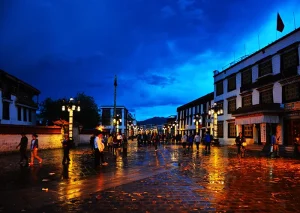 A vibrant night view of Barkhor Street with colorful lights reflecting on the wet pavement under a deep blue sky.