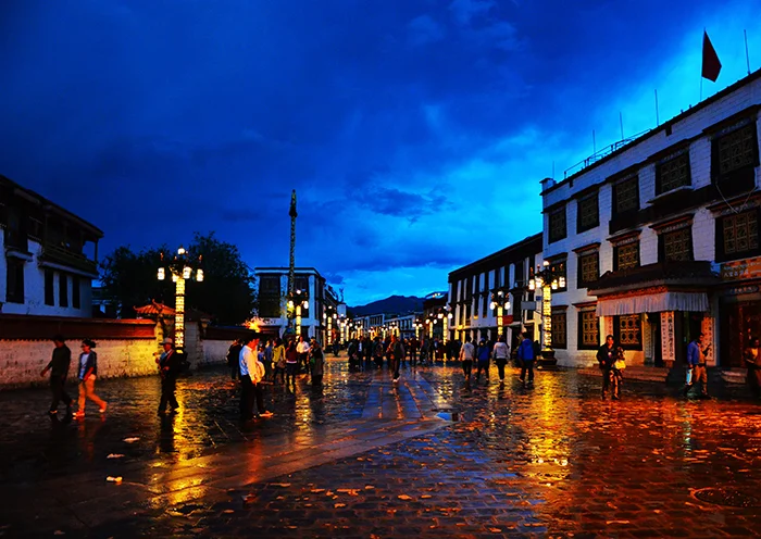 A vibrant night view of Barkhor Street with colorful lights reflecting on the wet pavement under a deep blue sky.