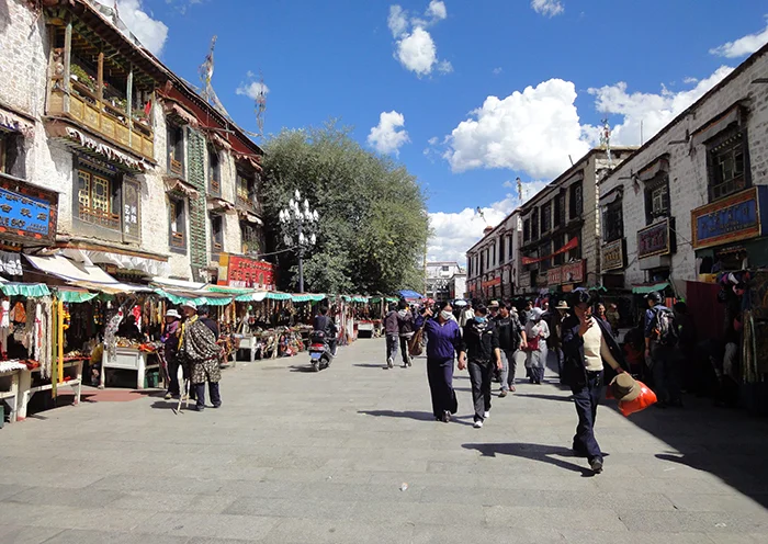 Crowds of tourists and locals walking past traditional market stalls on Barkhor Street under a cloudy blue sky.