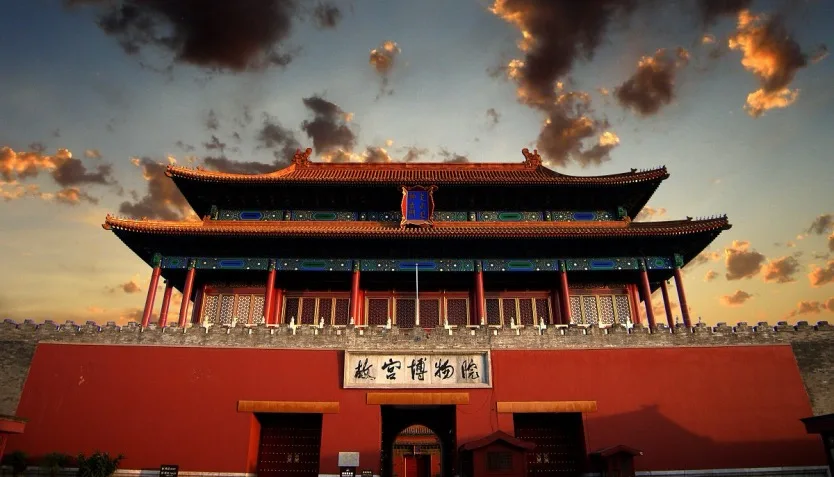 A low-angle shot of a grand traditional palace building inside the Forbidden City in Beijing, highlighting its intricate red pillars, golden glazed roof tiles, complex wooden brackets, and white marble terrace under a bright sky.