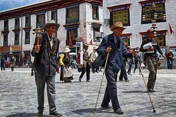 Elderly Tibetan pilgrims walking along the stone-paved Barkhor Street in Lhasa, holding prayer wheels and walking sticks against the backdrop of traditional white-washed buildings.
