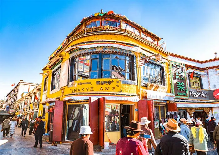 The bright yellow traditional Tibetan building of Makye Ame restaurant located on Barkhor Street in Lhasa, surrounded by tourists and pilgrims under a clear blue sky.