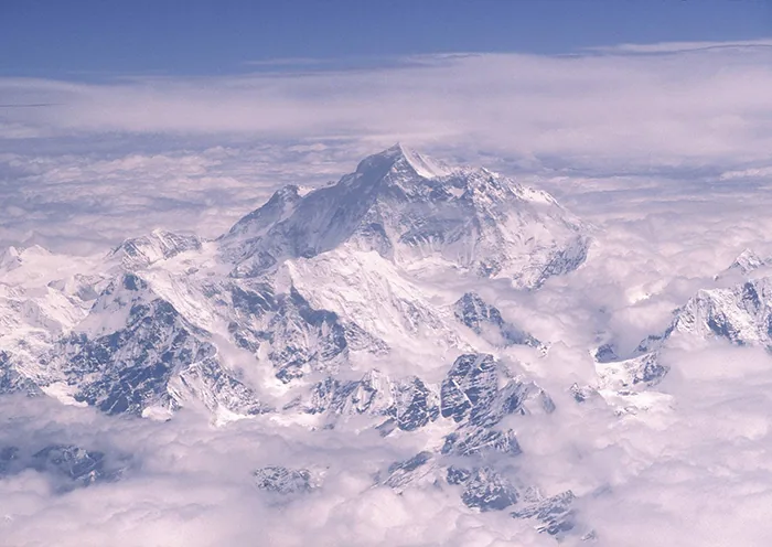 An aerial photograph of the snow-covered pyramidal peak of Mount Everest rising above a vast sea of white clouds in the Shigatse region of Tibet.