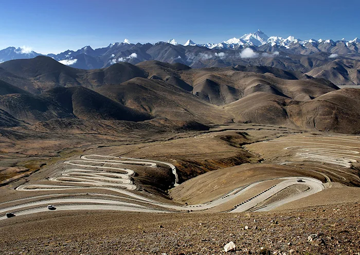 A long serpentine mountain road with sharp switchbacks winds through barren brown hills toward the snow-capped peak of Mount Everest in Shigatse, Tibet.