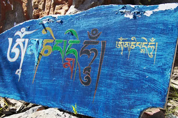 A close-up of a blue-painted stone inscribed with the sacred Tibetan mantra "Om Mani Padme Hum" in vibrant white, green, and red calligraphy.