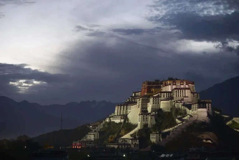 The Potala Palace captured from a distance under a dramatic cloudy sky, with dark mountains in the background.