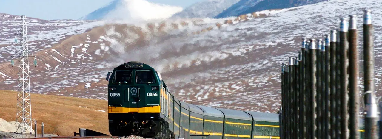 A green and yellow Qinghai-Tibet Railway train (Locomotive 0055) traveling through the permafrost region with cooling thermosyphons in the foreground and snow-capped mountains in the background.