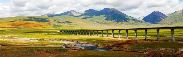A wide panoramic view of a long railway bridge spanning across the green Tibetan plateau with a backdrop of rolling mountains.