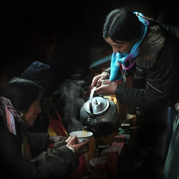A cinematic shot in a dimly lit environment showing a woman in a blue scarf pouring steaming hot sweet tea from a large kettle into a bowl held by another person.