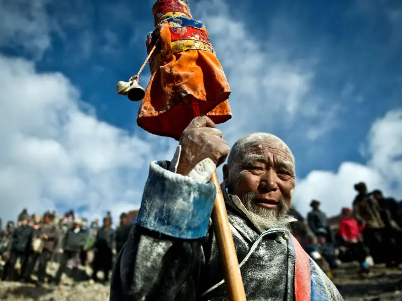 A close-up of a weathered Tibetan elder man with white powder on his face, holding a decorative spiritual item high against a bright blue sky during a crowded festival.