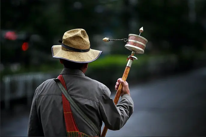 Rear view of a Tibetan man wearing a traditional hat and carrying a shoulder bag, holding a wooden handheld prayer wheel while walking in Lhasa.