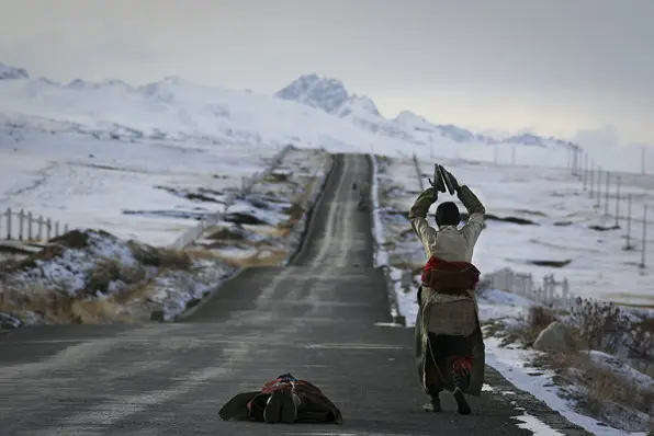 Two Tibetan pilgrims performing traditional full-body prostrations on a paved highway leading toward snow-capped mountains, a testament to their spiritual devotion during the long journey to Lhasa.