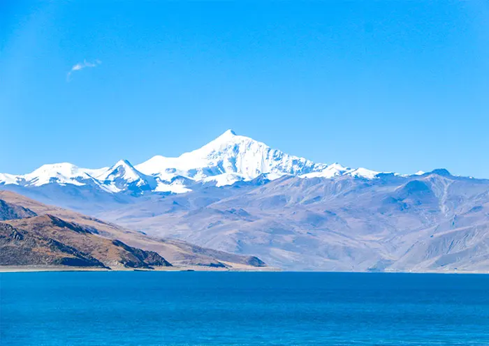 A high-altitude landscape photograph of the turquoise Yamdrok Lake in Tibet, featuring the snow-capped peak of Mount Nyenchen Khangsar against a clear blue sky.