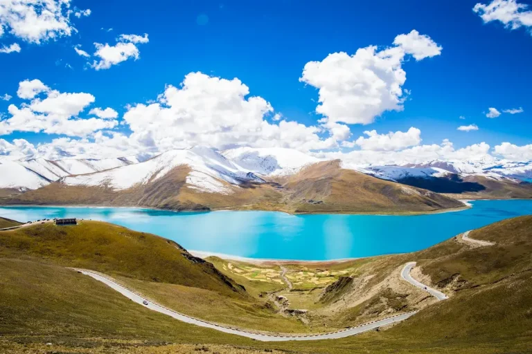 Panoramic view of the turquoise Yamdrok Lake in Tibet with a winding mountain road in the foreground.
